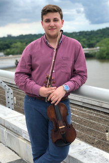 Photo of George Pekarsky, a young man, standing with legs crossed and leaning on a short wall. He is facing the camera and holding a violin.