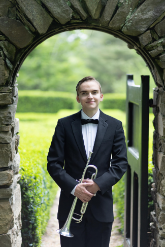 Photo of Levi Pardy, a smiling young man wearing a standard black tuxedo with a black bow tie and hold ing a trumpet. He is standing in a stone arch doorway, with a garden behind him.